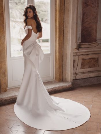 A woman wearing the TRISHA by Enzoani bridal gown—an off-the-shoulder white dress with bow and button accents—stands near a tall window in an ornate room.