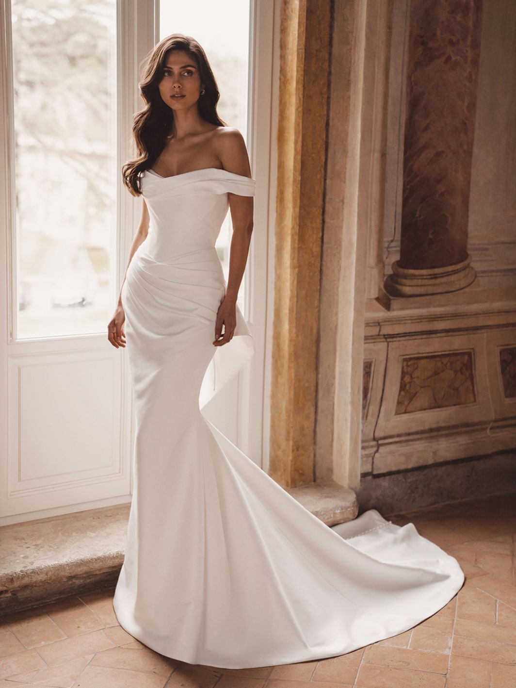 A woman stands indoors by a tall window and ornate wall, wearing the TRISHA by Enzoani wedding dress—an off-the-shoulder, fitted white gown with a long train. A woman stands indoors by a tall window and ornate wall, wearing the TRISHA by Enzoani wedding dress—an off-the-shoulder, fitted white gown with a long train.