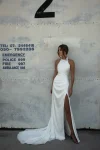 Woman in the WALSH white halter-neck dress with a thigh-high slit stands against a worn concrete wall marked with emergency numbers and a large black number 2.