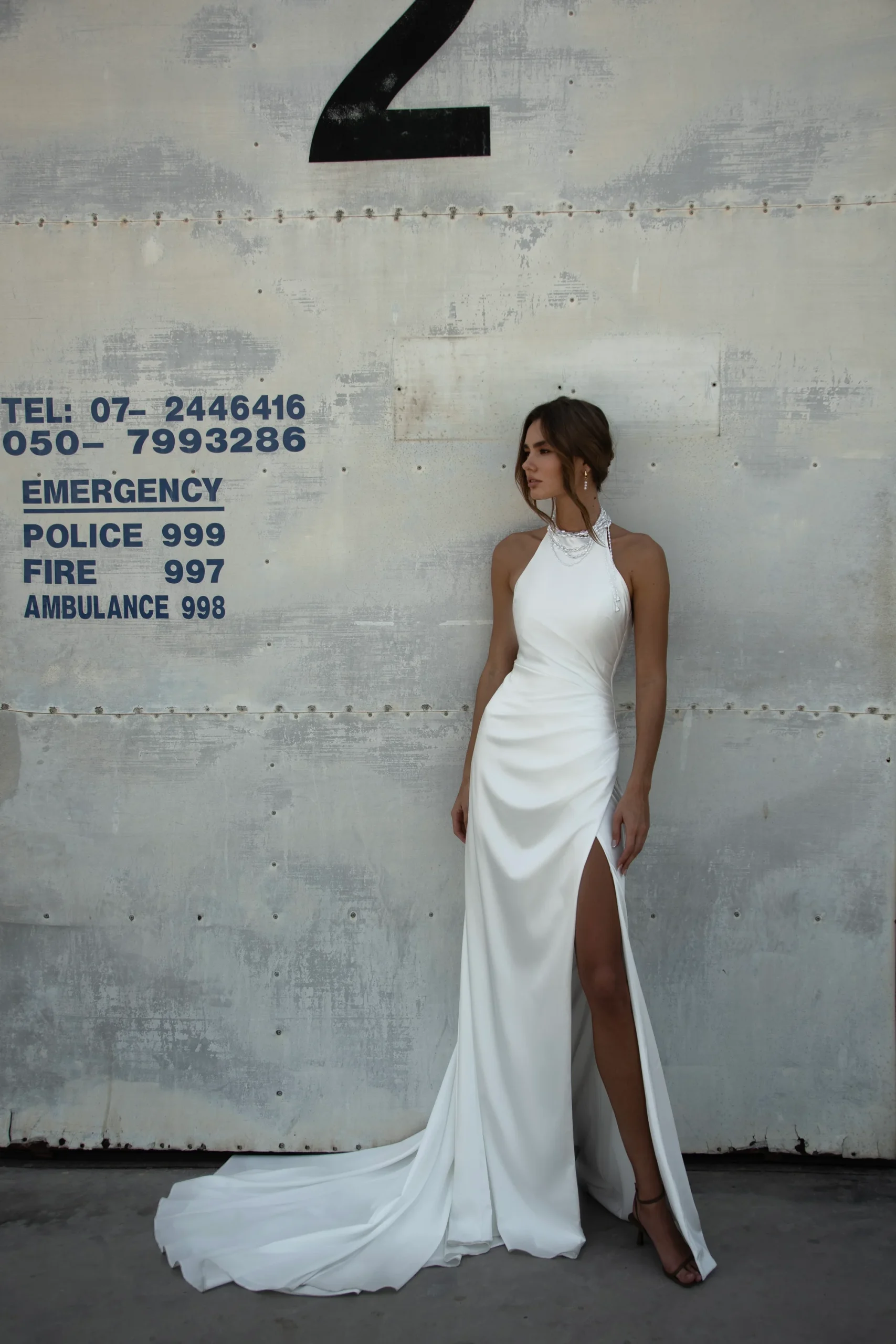 Woman in the WALSH white halter-neck dress with a thigh-high slit stands against a worn concrete wall marked with emergency numbers and a large black number 2. Woman in the WALSH white halter-neck dress with a thigh-high slit stands against a worn concrete wall marked with emergency numbers and a large black number 2.