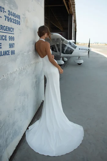 A woman in the WALSH backless white gown stands against a hangar wall near a small white aircraft on a concrete surface.