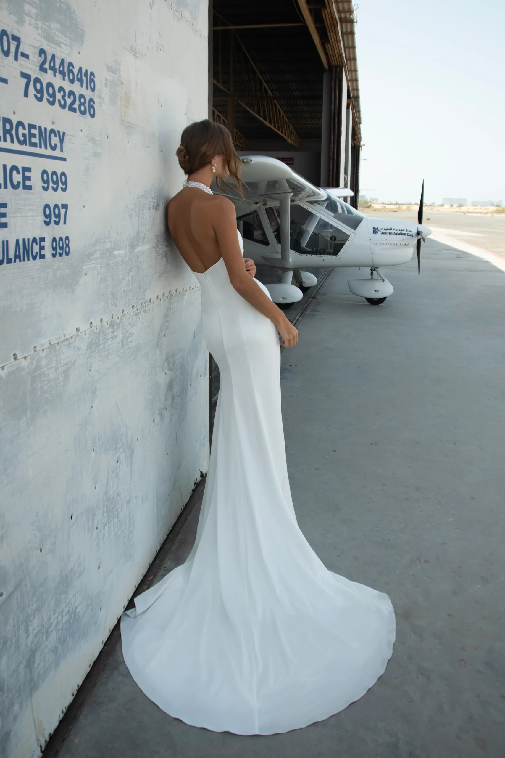 A woman in the WALSH backless white gown stands against a hangar wall near a small white aircraft on a concrete surface. A woman in the WALSH backless white gown stands against a hangar wall near a small white aircraft on a concrete surface.