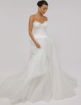 A woman wearing the CASSIA/AW2614 strapless white wedding gown with a fitted bodice and flowing tulle skirt stands against a plain light background.