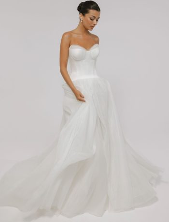 A woman wearing the CASSIA/AW2614 strapless white wedding gown with a fitted bodice and flowing tulle skirt stands against a plain light background.
