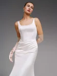 A woman stands in a studio wearing the DENNISE by Pronovias, a fitted white satin wedding dress featuring a sleeveless design, square neckline, and long train.
