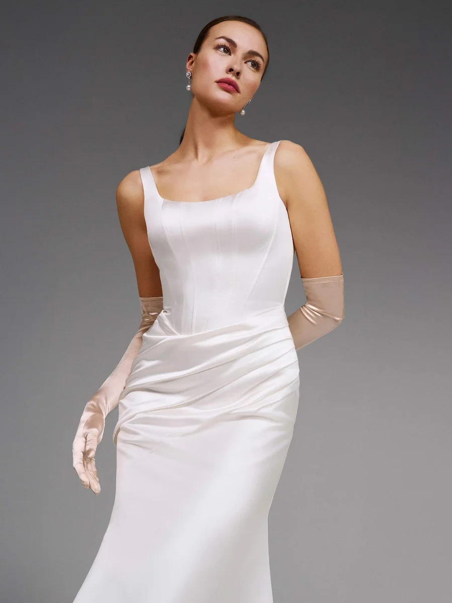 A woman in the DENNISE by Pronovias sleeveless white satin wedding dress with a square neckline and long pale gloves poses against a plain gray background. A woman in the DENNISE by Pronovias sleeveless white satin wedding dress with a square neckline and long pale gloves poses against a plain gray background.