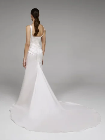 A woman faces away, wearing the DENNISE by Pronovias—a white sleeveless wedding gown with a fitted bodice, lace-up back, and long flowing train—against a plain gray background.