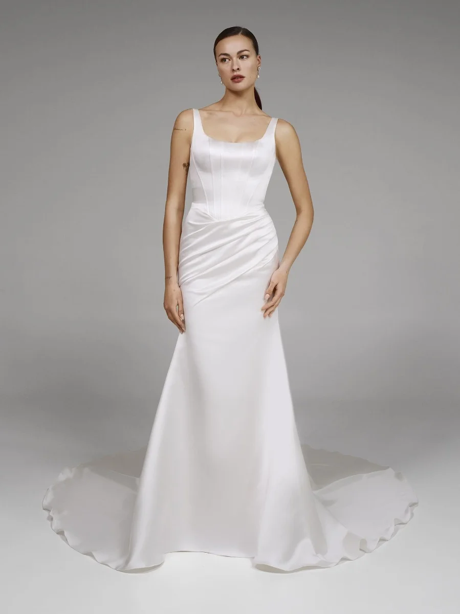 A woman stands in a studio wearing the DENNISE by Pronovias, a fitted white satin wedding dress featuring a sleeveless design, square neckline, and long train. A woman stands in a studio wearing the DENNISE by Pronovias, a fitted white satin wedding dress featuring a sleeveless design, square neckline, and long train.