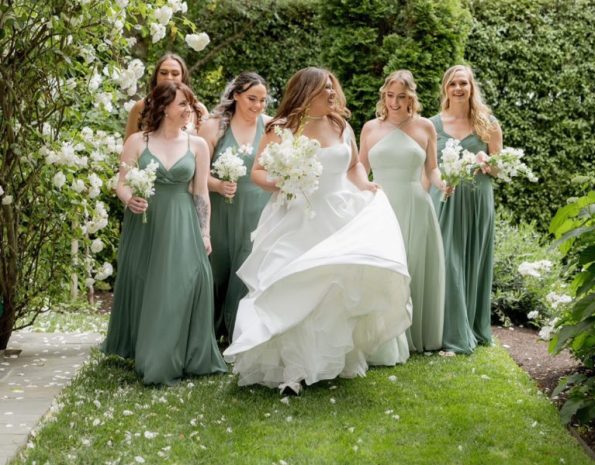 A bride in a white dress walks outdoors in Walnut Creek with five bridesmaids in sage green dresses, holding white bouquets, surrounded by greenery and white flowers.