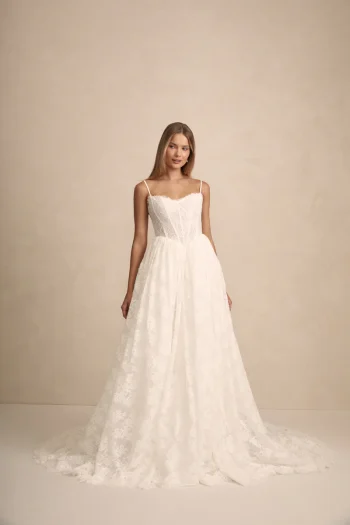 A woman wears the PRIM by Made with Love white lace wedding dress, featuring thin straps and a full, floor-length skirt, set against a plain beige background.