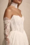 A woman in the strapless PRIM wedding dress by Made with Love stands outdoors in a courtyard, surrounded by greenery and elegant stone structures.