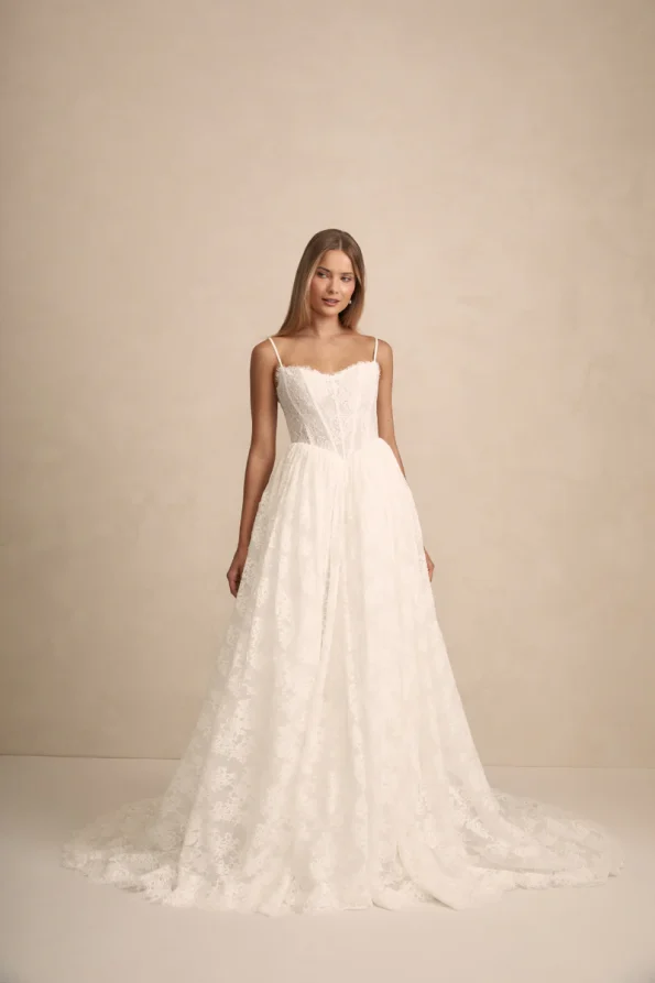 A woman wears the PRIM by Made with Love white lace wedding dress, featuring thin straps and a full, floor-length skirt, set against a plain beige background.