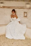A woman in the strapless PRIM wedding dress by Made with Love stands outdoors in a courtyard, surrounded by greenery and elegant stone structures.