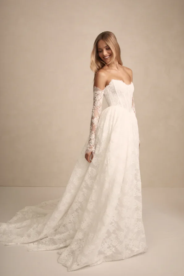 A woman smiles and looks down while wearing the PRIM strapless white lace wedding gown by Made with Love, with matching lace sleeves, against a plain beige background.