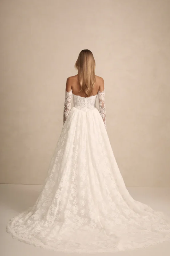 A woman with long blonde hair wears the PRIM by Made with Love off-the-shoulder white lace wedding gown with a long train, captured from behind against a neutral background, showcasing the brand’s signature elegance.