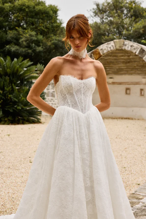 A woman in the strapless PRIM wedding dress by Made with Love stands outdoors in a courtyard, surrounded by greenery and elegant stone structures.