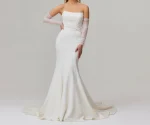 Wearing the TINA by Jessica Couture strapless, fitted white mermaid bridal gown with a long train and sheer gloves, a woman stands against a plain background.