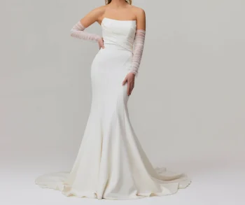 Wearing the TINA by Jessica Couture strapless, fitted white mermaid bridal gown with a long train and sheer gloves, a woman stands against a plain background.