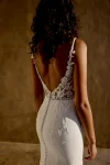 A woman with curly hair stands indoors near a large ceramic vase against a brown wall, wearing the sleeveless white 66447 wedding dress by Lillian West, featuring intricate lace details.