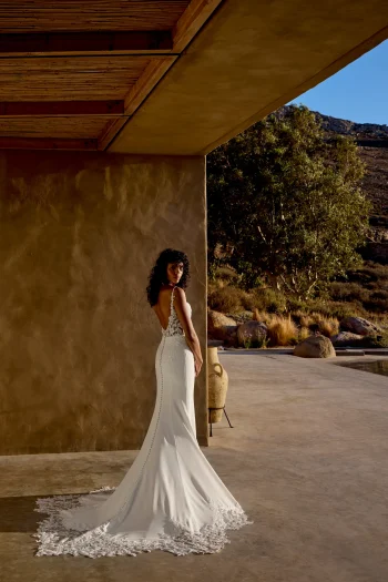 A woman in a white, backless 66447 by Lillian West wedding dress stands outside near a stucco wall, looking over her shoulder, with dry grass and hills in the background.