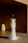 A woman with curly hair stands indoors near a large ceramic vase against a brown wall, wearing the sleeveless white 66447 wedding dress by Lillian West, featuring intricate lace details.