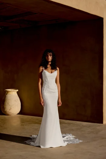 A woman with curly hair stands indoors near a large ceramic vase against a brown wall, wearing the sleeveless white 66447 wedding dress by Lillian West, featuring intricate lace details.