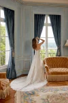 A woman wearing the 88525/MERIDA by Justin A—a strapless white lace wedding gown—stands in a sunlit, elegant room with blue curtains, wooden floors, and large windows, capturing the timeless romance and refined artistry that define Justin A’s vision.