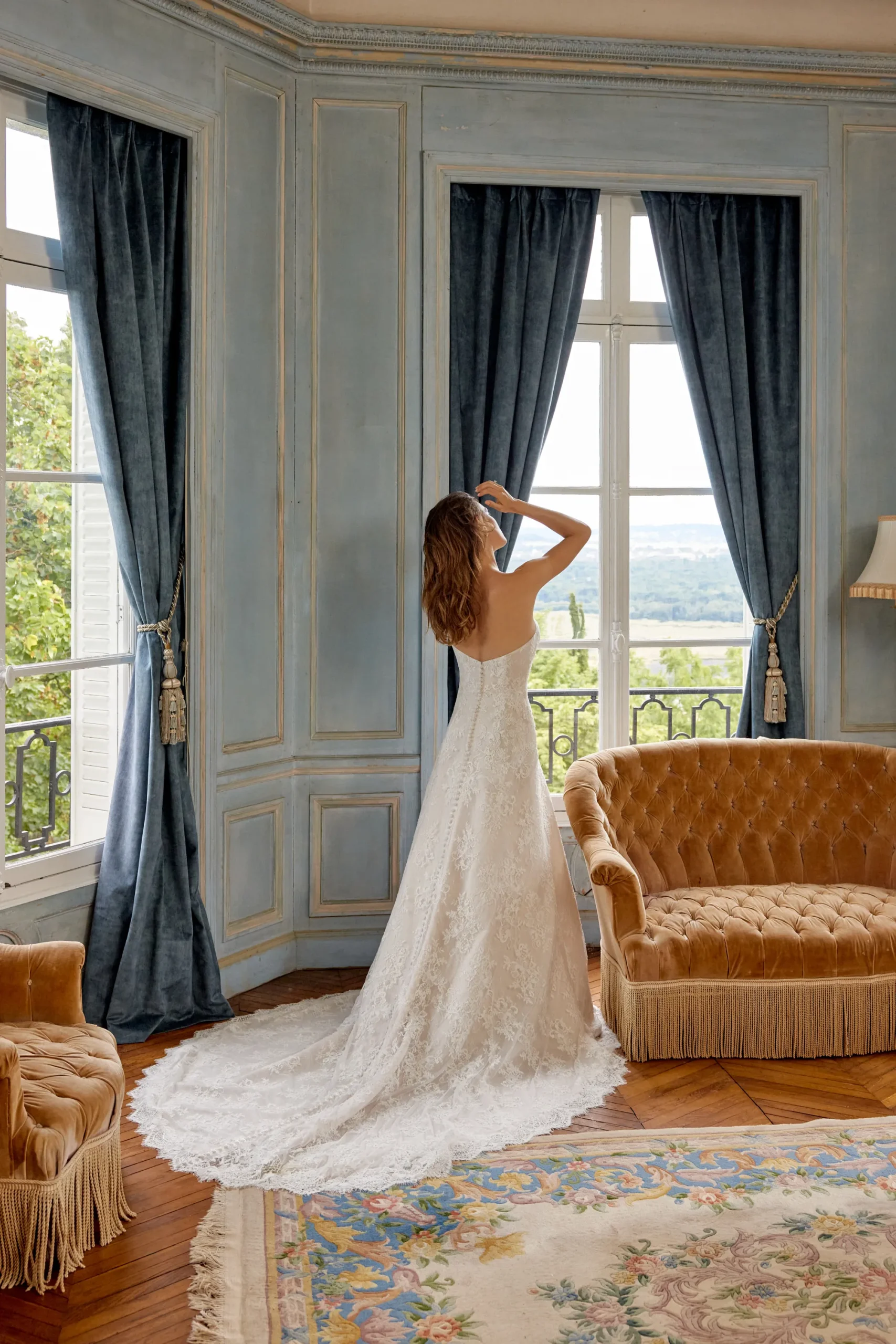 A woman wearing the 88525/MERIDA strapless white wedding dress by Justin Alexander stands in front of tall windows with dark blue curtains in an elegant room with gold sofas. A woman wearing the 88525/MERIDA strapless white wedding dress by Justin Alexander stands in front of tall windows with dark blue curtains in an elegant room with gold sofas.