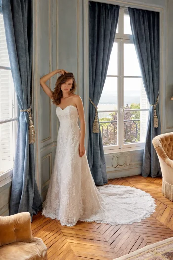 A woman wearing the 88525/MERIDA by Justin A—a strapless white lace wedding gown—stands in a sunlit, elegant room with blue curtains, wooden floors, and large windows, capturing the timeless romance and refined artistry that define Justin A’s vision.