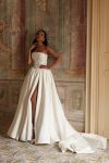A woman showcases the ARABELLA gown—a strapless white dress featuring a beaded bodice and high slit—while posing indoors against an ornate wall and tiled floor, standing near a beige door.