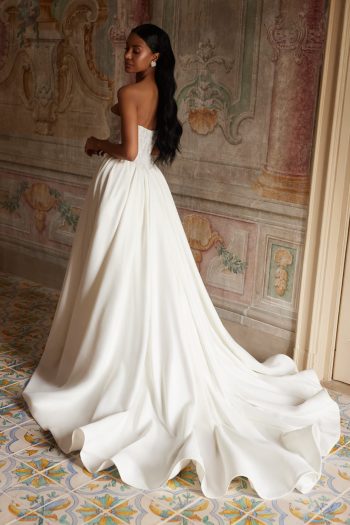 A woman wearing the ARABELLA strapless white wedding gown with a long train stands on a patterned floor in an ornate, vintage-style room.