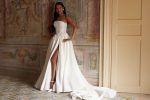 A woman showcases the ARABELLA gown—a strapless white dress featuring a beaded bodice and high slit—while posing indoors against an ornate wall and tiled floor, standing near a beige door.