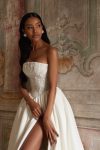 A woman showcases the ARABELLA gown—a strapless white dress featuring a beaded bodice and high slit—while posing indoors against an ornate wall and tiled floor, standing near a beige door.