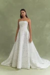 A woman stands against a green background wearing the BRODY by Jenny Yoo, a strapless, floor-length white gown with a floral textured pattern.