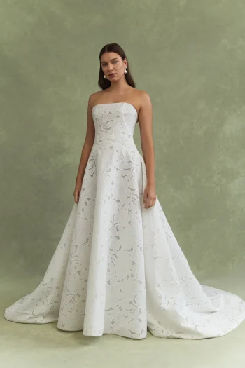A woman stands against a green background wearing the BRODY by Jenny Yoo, a strapless, floor-length white gown with a floral textured pattern.