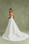 A woman stands against a green background wearing the BRODY by Jenny Yoo, a strapless, floor-length white gown with a floral textured pattern.