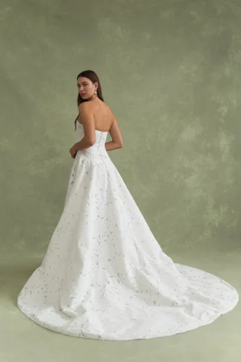 A woman in the strapless white BRODY by Jenny Yoo gown with a long train stands against a textured green background, looking over her shoulder.