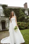A woman stands against a green background wearing the BRODY by Jenny Yoo, a strapless, floor-length white gown with a floral textured pattern.