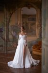 A woman wearing the CAPELLA, an elegant off-the-shoulder white wedding dress with lace sleeves and a thigh-high slit, stands in an ornately decorated room with painted walls and a large mirror.
