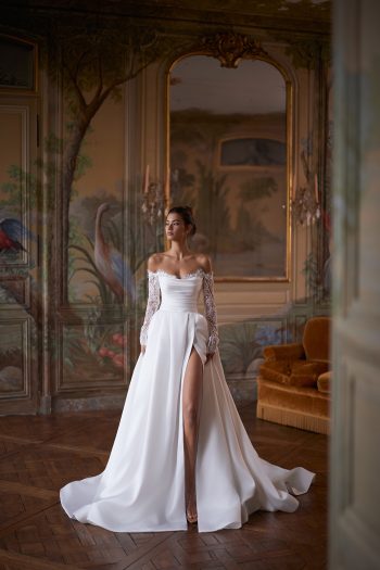 A woman wearing the CAPELLA, an elegant off-the-shoulder white wedding dress with lace sleeves and a thigh-high slit, stands in an ornately decorated room with painted walls and a large mirror.