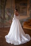 A woman wearing the CAPELLA, an elegant off-the-shoulder white wedding dress with lace sleeves and a thigh-high slit, stands in an ornately decorated room with painted walls and a large mirror.