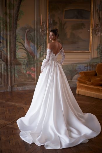 Wearing the CAPELLA gown—an off-the-shoulder white wedding dress with elegant lace sleeves—a woman stands in an ornate room featuring a painted mural and wooden floors, glancing over her shoulder.