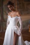 A woman wearing the CAPELLA, an elegant off-the-shoulder white wedding dress with lace sleeves and a thigh-high slit, stands in an ornately decorated room with painted walls and a large mirror.