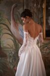 A woman wearing the CAPELLA, an elegant off-the-shoulder white wedding dress with lace sleeves and a thigh-high slit, stands in an ornately decorated room with painted walls and a large mirror.