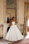 Bridal Description: The DELPHINA by Nava Bride woman in a strapless white ball gown stands gracefully in front of a fireplace with an ornate mirror and classical decor, embodying timeless elegance in this sophisticated setting.