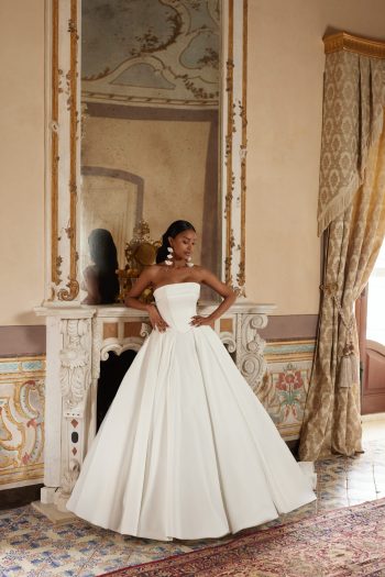 Bridal Description: The DELPHINA by Nava Bride woman in a strapless white ball gown stands gracefully in front of a fireplace with an ornate mirror and classical decor, embodying timeless elegance in this sophisticated setting.