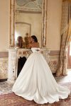 Bridal Description: The DELPHINA by Nava Bride woman in a strapless white ball gown stands gracefully in front of a fireplace with an ornate mirror and classical decor, embodying timeless elegance in this sophisticated setting.