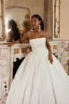 Bridal Description: The DELPHINA by Nava Bride woman in a strapless white ball gown stands gracefully in front of a fireplace with an ornate mirror and classical decor, embodying timeless elegance in this sophisticated setting.