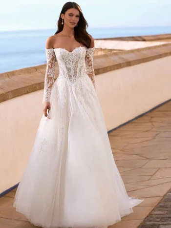 A woman stands outdoors near a stone railing, framed by the ocean and sky, wearing the stunning FAIZA gown by Love by Enz—a long-sleeved, off-the-shoulder white lace wedding dress with a voluminous full skirt.