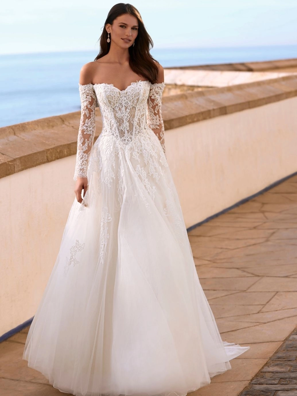 A woman stands outdoors near a stone railing, framed by the ocean and sky, wearing the stunning FAIZA gown by Love by Enz—a long-sleeved, off-the-shoulder white lace wedding dress with a voluminous full skirt. A woman stands outdoors near a stone railing, framed by the ocean and sky, wearing the stunning FAIZA gown by Love by Enz—a long-sleeved, off-the-shoulder white lace wedding dress with a voluminous full skirt.